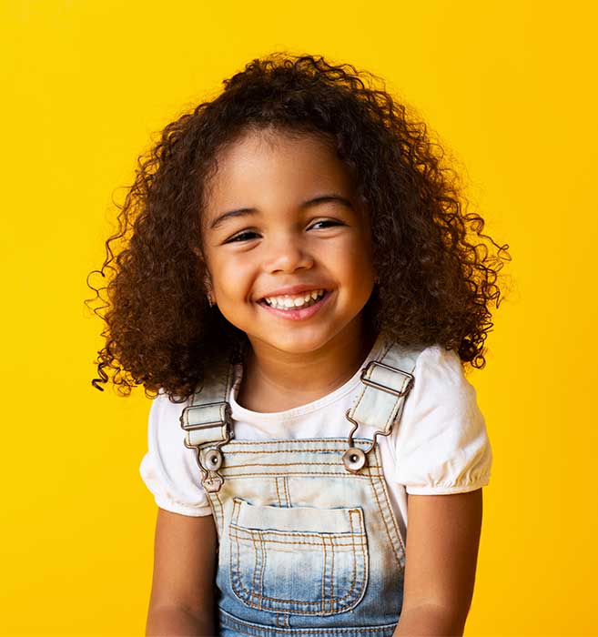 Young girl smiling at camera wearing overalls against bright yellow background.