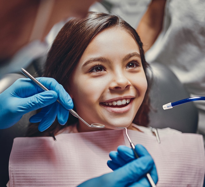 Young girl smiling at dental checkup with dentist holding tools wearing blue gloves.
