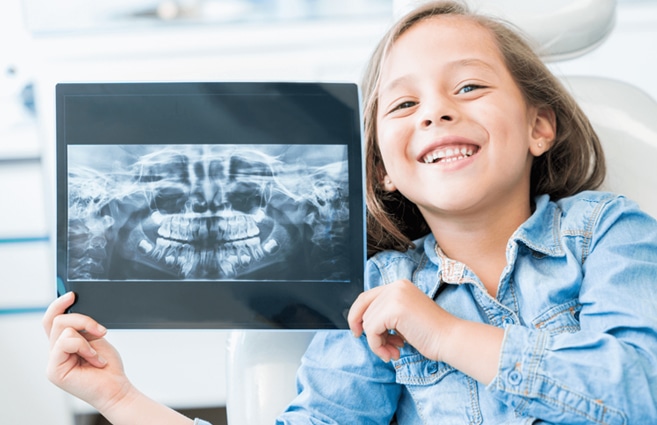 Young girl smiling while holding dental X-ray in modern dental office setting.