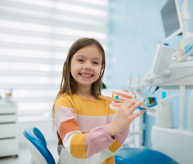 Young girl smiling in dental office chair holding toothbrush near modern dental equipment.