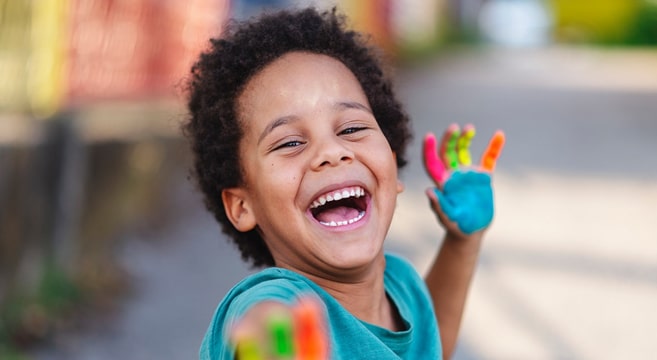 Happy child with colorful painted hand smiling outdoors in bright daylight setting.