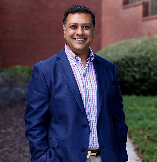 Professional headshot of smiling man in blue blazer and checkered shirt outdoors against brick building.
