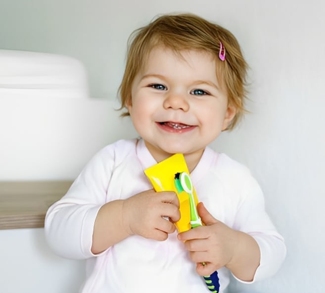Smiling toddler in white shirt holding yellow toothbrush in bright dental office setting.