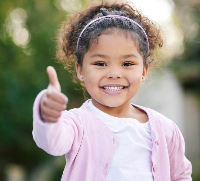 Young child smiling brightly with thumbs up gesture outdoors in natural setting.