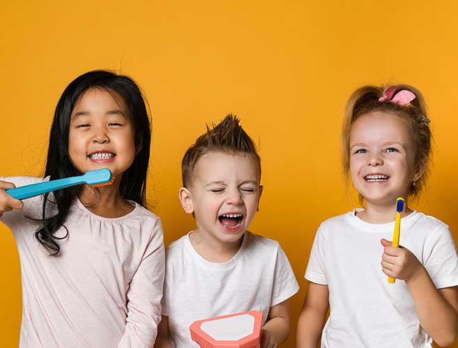 Three happy children holding toothbrushes against yellow background for dental care.
